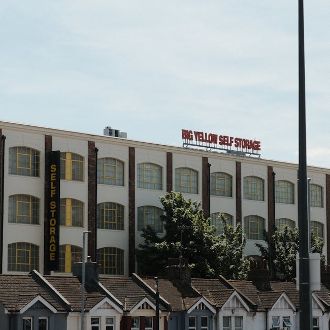 A multi-storey commercial building with a white facade and large, evenly spaced yellow-framed windows. The building has a vertical yellow sign on the left side that reads 'SELF STORAGE' and a large re